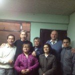 Group of seven people posing indoors, wearing casual jackets, with a neutral background.