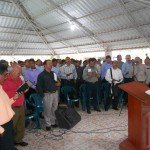 Group of people singing at church gathering under a metal roof, led by a speaker at a podium.