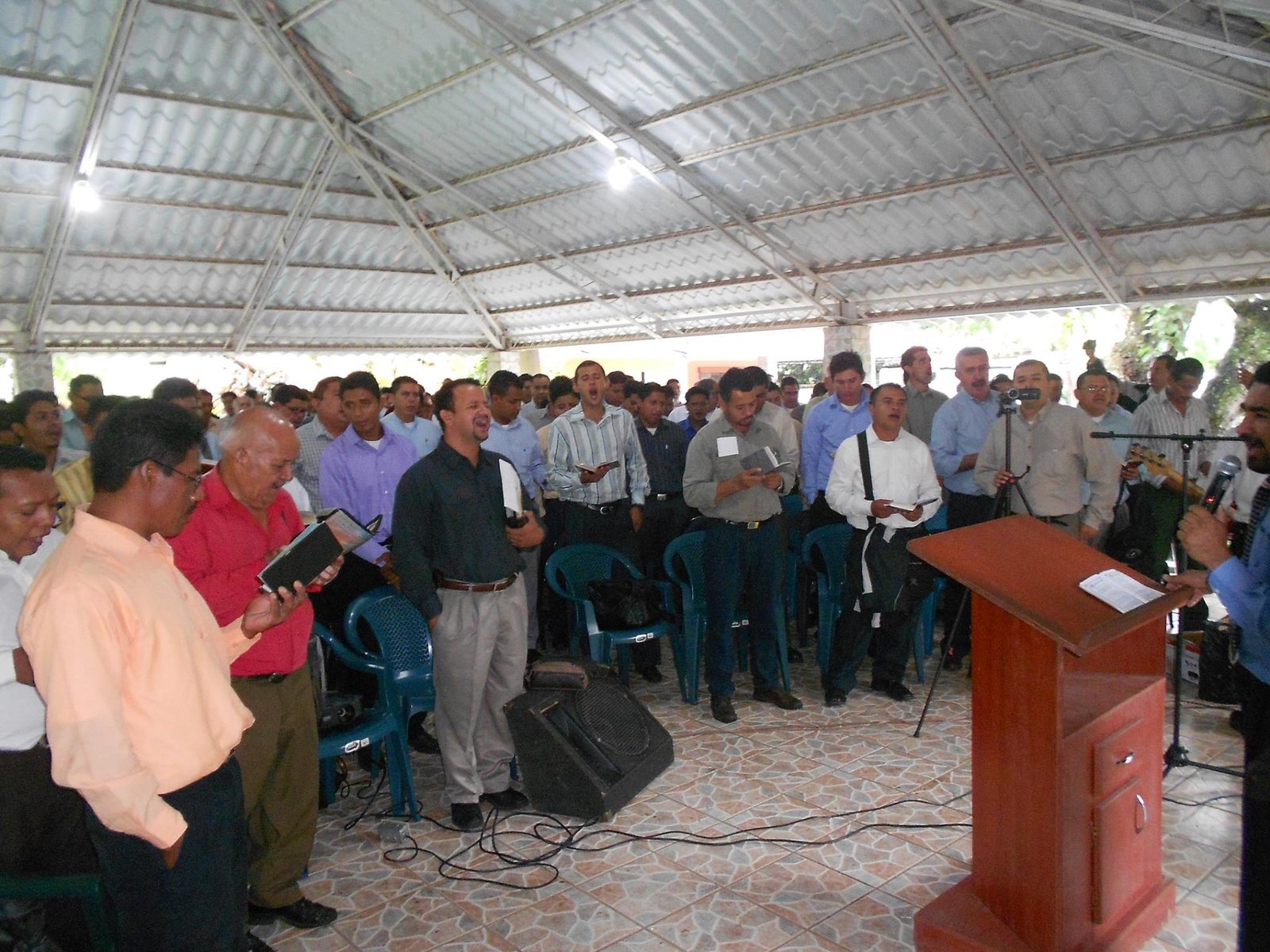 Group of people singing at church gathering under a metal roof, led by a speaker at a podium.