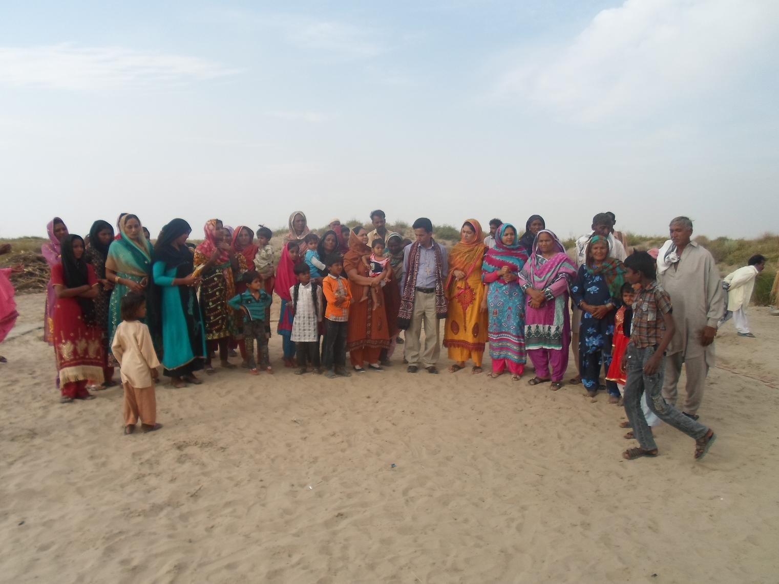 Group of people standing together on a sandy beach, wearing colorful traditional attire under a partly cloudy sky.