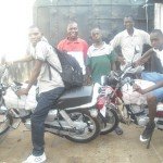 Group of men with motorcycles, smiling outside a gated area, showcasing bikes and casual attire.
