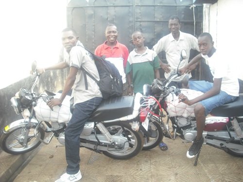 Group of men with motorcycles, smiling outside a gated area, showcasing bikes and casual attire.