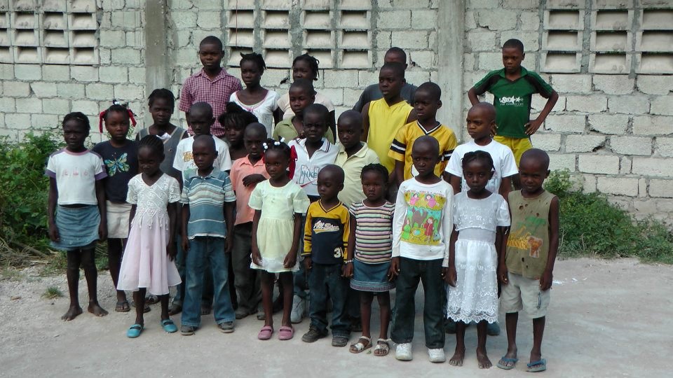 Group of children posing outside, standing in front of a brick wall, some wearing colorful outfits and smiling.