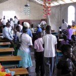 People gathered in a community hall with benches, decorated with hanging flowers and filled with natural light.