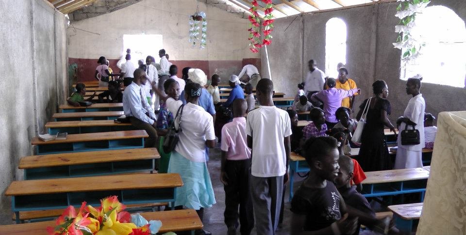 People gathered in a community hall with benches, decorated with hanging flowers and filled with natural light.