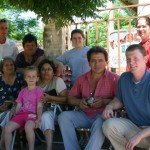 Group of people sitting and standing outdoors under a tree, smiling at the camera.
