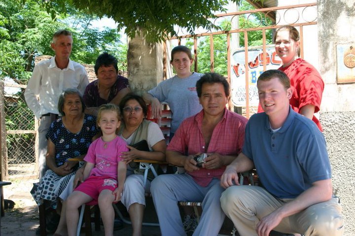 Group of people sitting and standing outdoors under a tree, smiling at the camera.
