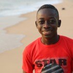 Smiling boy in red T-shirt on a sandy beach with ocean waves in the background.