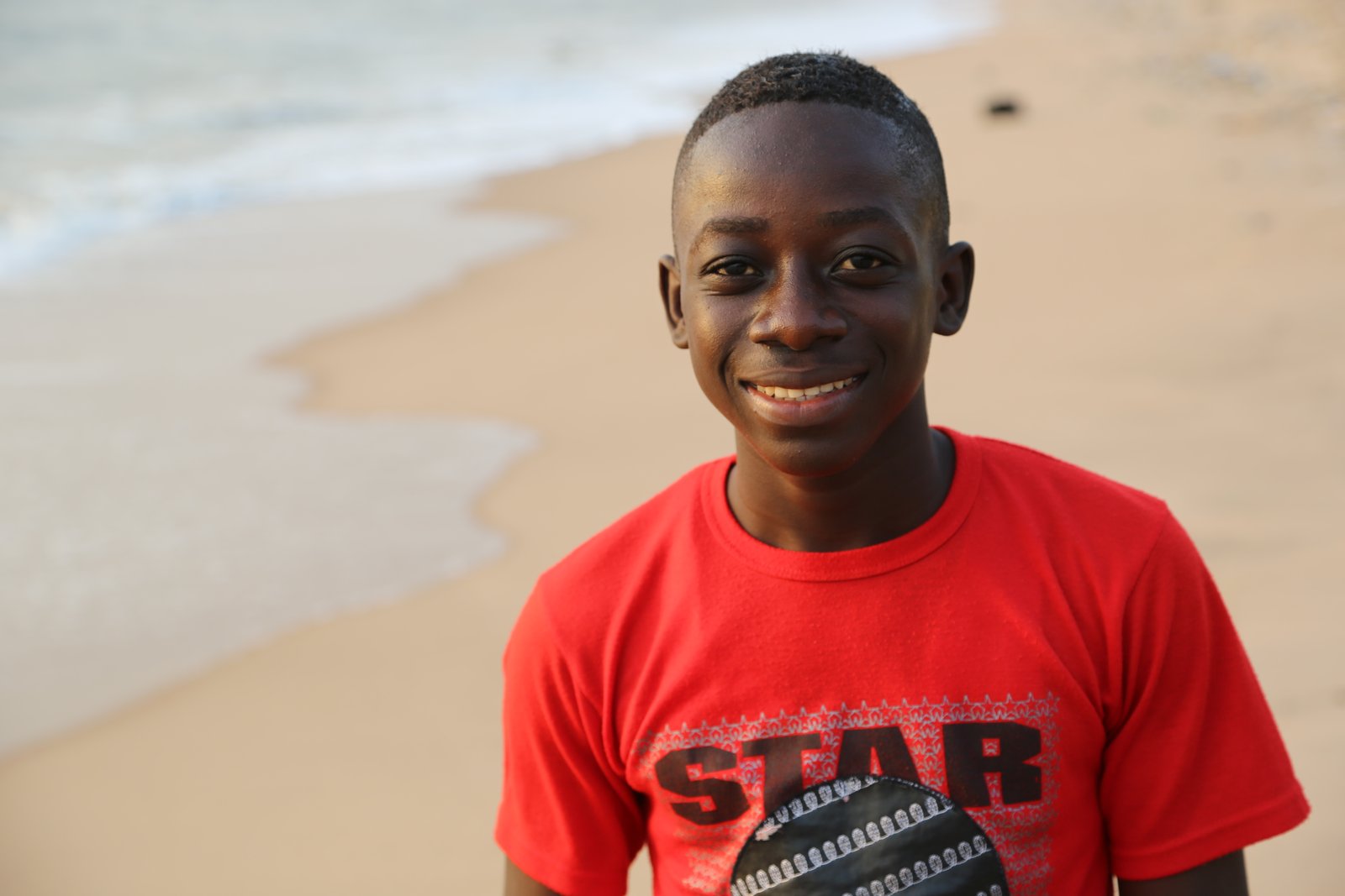 Smiling boy in red T-shirt on a sandy beach with ocean waves in the background.