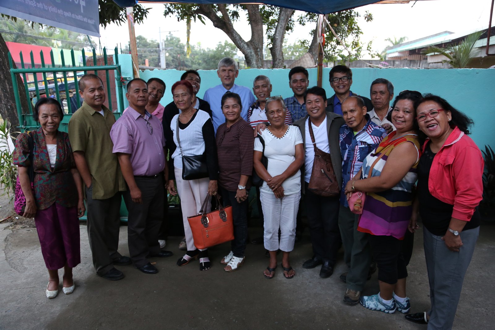 Group of people smiling and posing outdoors, with colorful buildings and trees in the background.