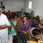 A group of people gathered in a room for prayer, some kneeling with hands folded, women in colorful saris.