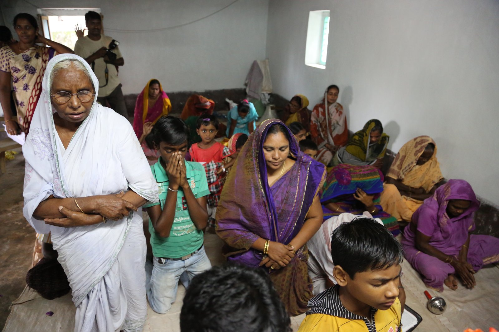 A group of people gathered in a room for prayer, some kneeling with hands folded, women in colorful saris.