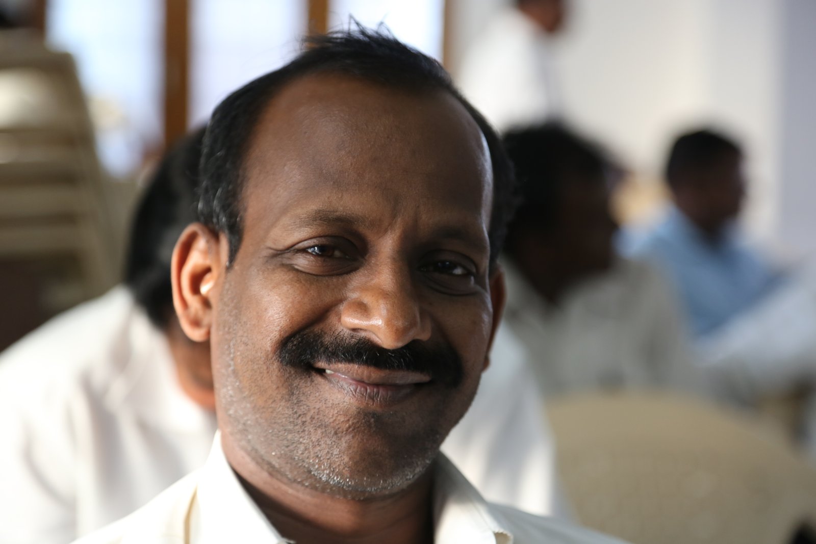Smiling man in a white shirt at an indoor gathering, creating a warm and inviting atmosphere.