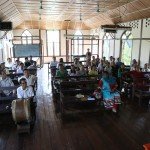 People attending a community meeting in a wooden, sunlit hall with high ceilings and fans.