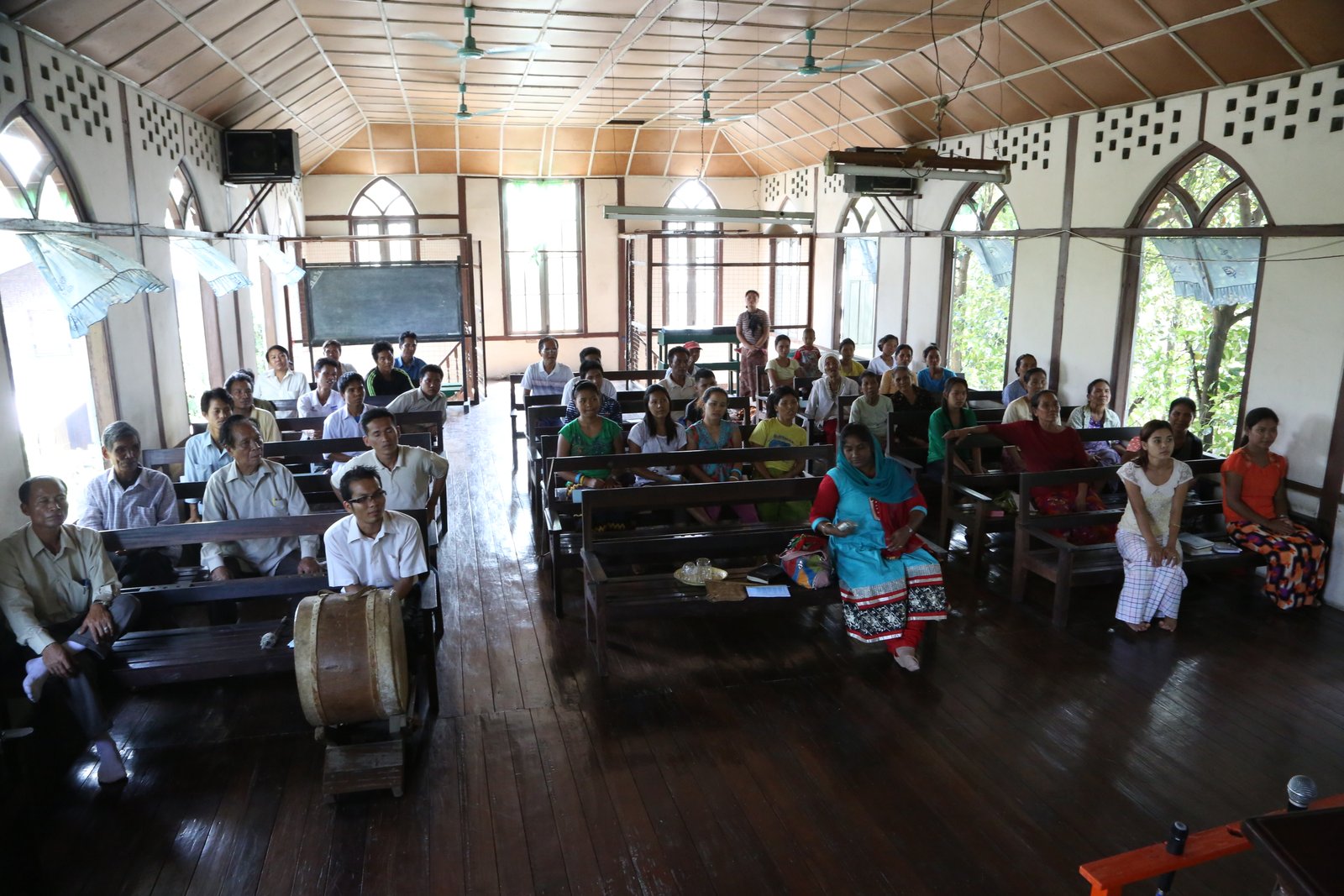People attending a community meeting in a wooden, sunlit hall with high ceilings and fans.