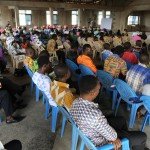 People attending a community meeting indoors, seated on blue chairs, and listening attentively to a speaker.