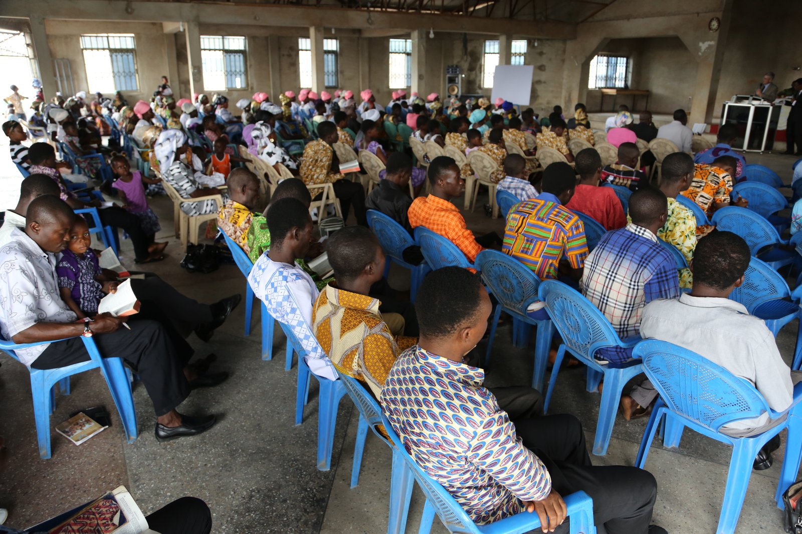 People attending a community meeting indoors, seated on blue chairs, and listening attentively to a speaker.