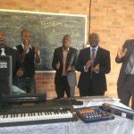 Men in suits clapping near musical equipment in a room with a chalkboard, promoting lively community engagement.