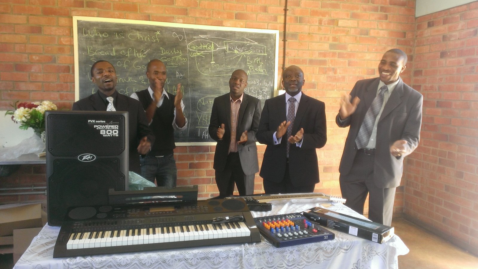 Men in suits clapping near musical equipment in a room with a chalkboard, promoting lively community engagement.