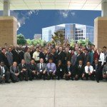 Group photo of diverse professionals in suits gathered outside a modern building with flags and a clear sky.