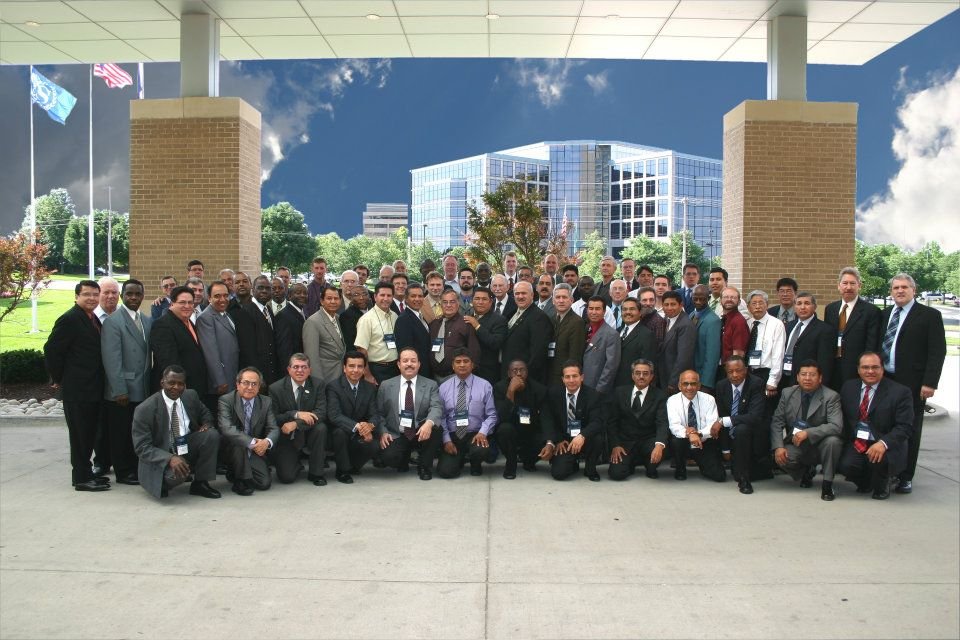 Group photo of diverse professionals in suits gathered outside a modern building with flags and a clear sky.