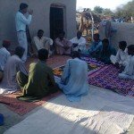 Group of men seated in a circle for a community meeting outdoors on colorful rugs near a mud house.