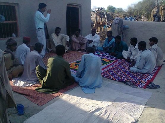Group of men seated in a circle for a community meeting outdoors on colorful rugs near a mud house.