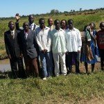 Group of people standing by a small lake in a rural landscape, with cattle in the background.