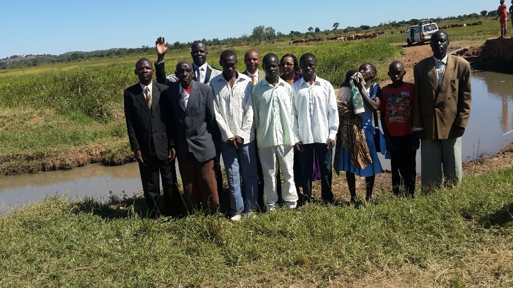 Group of people standing by a small lake in a rural landscape, with cattle in the background.