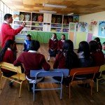 Teacher leading a group discussion with students seated in a classroom, colorful posters on the walls.