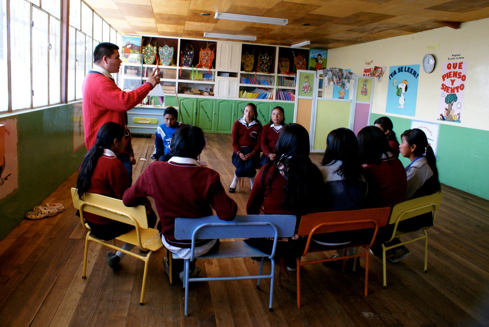Teacher leading a group discussion with students seated in a classroom, colorful posters on the walls.