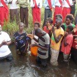 Group gathered in water for a baptism ceremony, some wearing colorful clothes, clapping and praying.