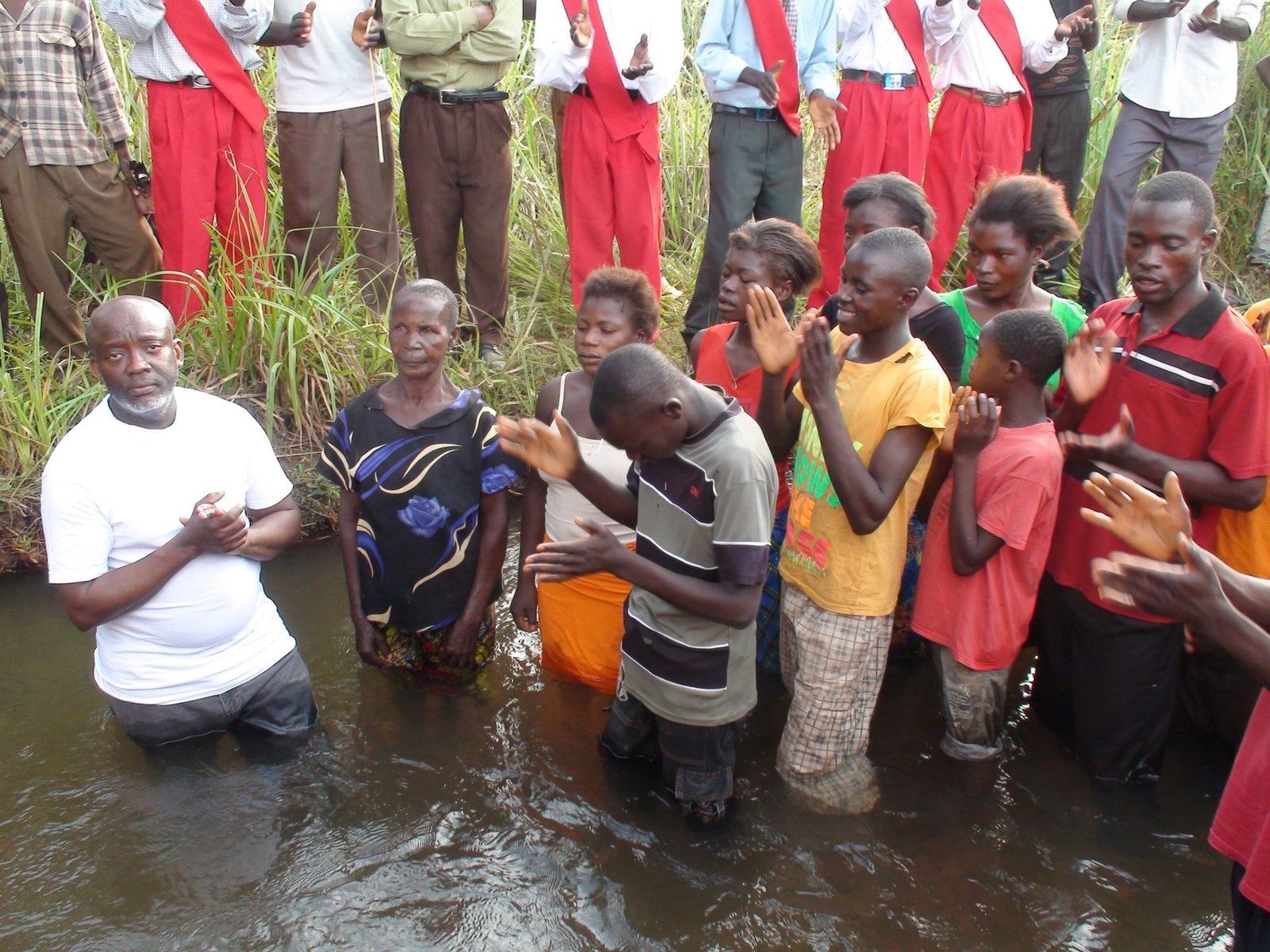 Group gathered in water for a baptism ceremony, some wearing colorful clothes, clapping and praying.