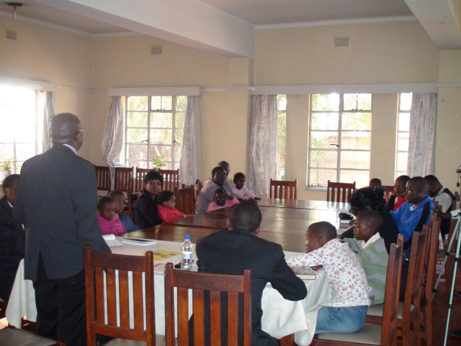 People attending a meeting in a room with large windows and wooden chairs.