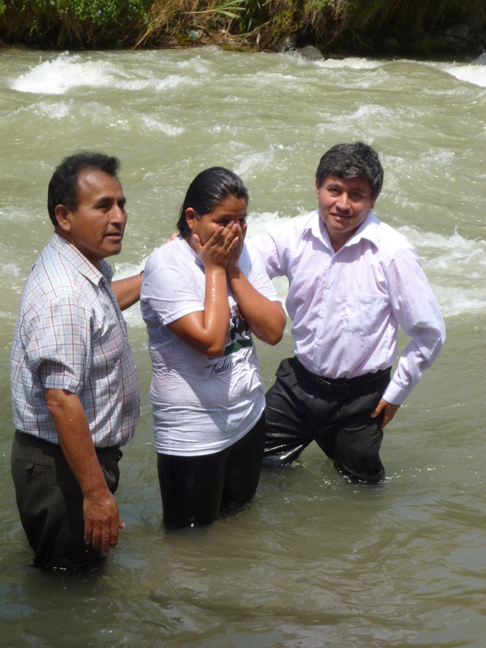 Three people standing in a river participating in an outdoor baptism ceremony.