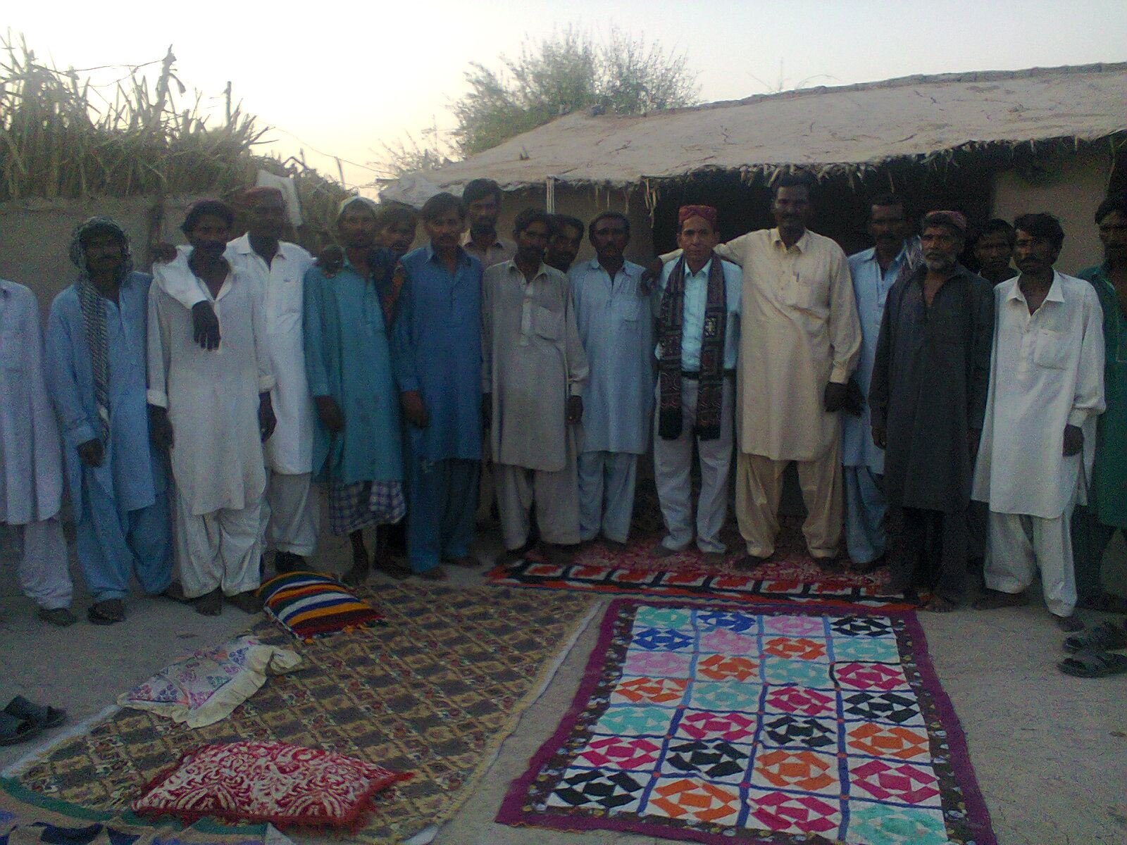 Group of men standing together in traditional attire in front of a rural hut with colorful rugs on the ground.