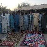 Group of men in traditional attire standing outdoors near a thatched structure with colorful blankets on the ground.