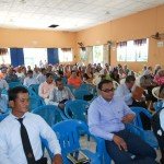 Audience seated at a community event in a hall, with men and women attentively listening.