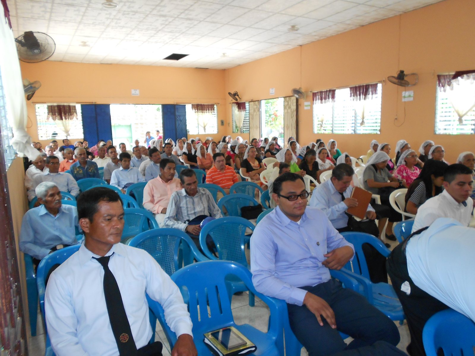 Audience seated at a community event in a hall, with men and women attentively listening.