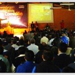 Large audience attending a presentation in a conference hall with a speaker on stage and projected displays.