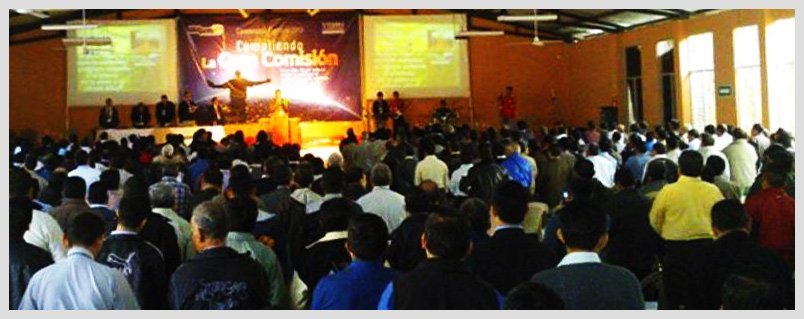 Large audience attending a presentation in a conference hall with a speaker on stage and projected displays.