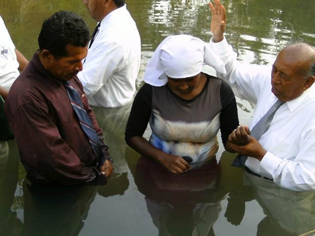 Religious baptism ceremony in a river with three people, two men and one woman, engaged in spiritual ritual.