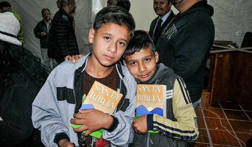 Two boys holding Bibles at a community gathering, surrounded by people inside a building.