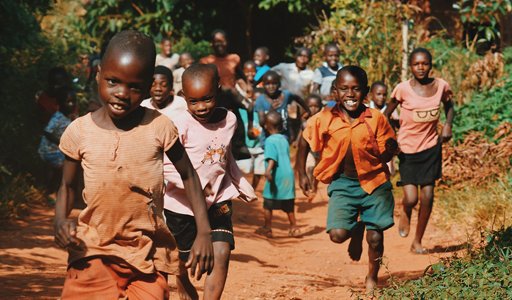 Group of children joyfully running on a dirt path in a rural setting, surrounded by green foliage.