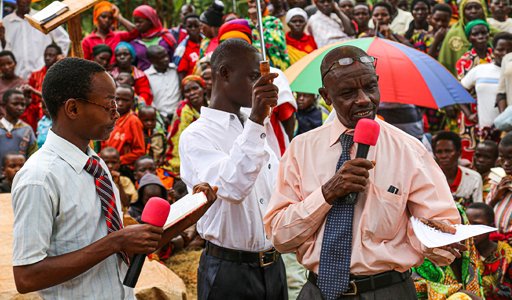 Community gathering with speakers under colorful umbrella, engaging audience with microphones and notes.